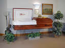 Dearly Departed - photo of a red casket in a room. The casket is open for viewing. Flowers and wreaths are placed around it.