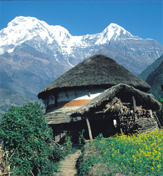 Nepal - Its a Nepal house sitting off a dirt road. In the background is a mountain with snow on the top of it and towards the front of the picture are green weeds.