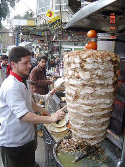 Shawarma - Shawarma is made by placing strips of meat or marinated chicken on a skewer. Animal fat and an onion or tomato are placed at the top of the stack to provide flavoring. The meat is then roasted slowly on all sides as the skewer rotates in front of or over a flame for a period of several hours (see rotisserie). Traditionally a wood fire is used, but recently a gas flame is more common. While many specialty restaurants might offer two or more, usually of different meats some establishments have one skewer.  A cheap version of shawarma is made of ground meat that is pre-formed and frozen onto a skewer. This is the most popular form of shawarma in Finland and Southern California but almost unheard of in Central Europe.  After cooking, the meat is shaved off the skewer with a large knife, an electric knife or a small circular saw, dropping to a circular tray below to be retrieved. Shawarma is most commonly eaten as a fast food, made up into a sandwich with pita bread or rolled up in lafa (a sweet, fluffy flatbread) together with vegetables and a dressing. Vegetables commonly found in shawarma include cucumber, onion, tomato, lettuce, parsley, pickled turnips, pickled gherkins, cabbage, and in some countries, such as Jordan or Saudi Arabia, french fries.  Common dressings include tahini (or tahina), Amba sauce (pickled mango with Chilbeh) and hummus, flavored with vinegar and spices such as cardamom, cinnamon, and nutmeg. Chicken shawarma is often served with garlic mayonnaise, pomegranate concentrate, skhug (a hot chili sauce), or any combination of the three. Once the sandwich is made, it might be dipped in the fat dripping from the skewer and then briefly seared against the flame. In Syria and Lebanon, chicken shawarma sandwiches are generally toasted after being made up, whereas those made of lamb or beef are immediately eaten.  Sometimes beef is used for shawarma instead of lamb, and turkey is also occasionally used instead of chicken. In Saudi Arabia, goat is equally as common as beef or lamb and is often the preferred primary meat for purists. In Israel, a turkey/lamb fat mixture is the primary flavor, although chicken is also available. Less common alternatives include fish and sausage. Some shawarma stores use hot dog buns or baguettes, but most have pita and lafa. Shawarma is often served with a plate of french fries or home fries. Sometimes, beef shawarma—despite its name—contains some lamb in addition to the beef, to ensure juiciness.  Shawarma is eaten either as a dish by itself, with grilled bread and a garnish, or with other Middle Eastern foods like Tabouli, Hummus, and Fattoush.