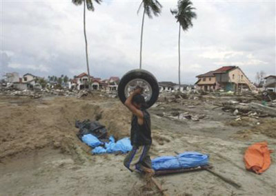 Images of Aceh 7 - Survivor walks past mass graves & bodies waiting burial with car wheel