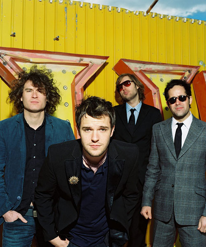 The Killers - Group shot Brandom kneeling into camera fram cneter yellow bright backdrop frames the band.