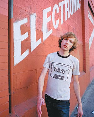 Beck - Full shot BH wearing a white t-shirt against a orage brick backdrop