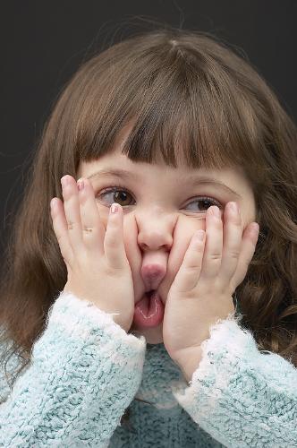 Child being silly - This is a photograph of a young child with brown hair. She has large eyes and is wearing a wooly sweater or cardigan. The has her hands up on her face and is pushing her cheeks together. It's a very silly picture of a little girl.