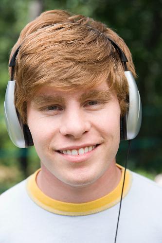Listening to Music - Picture of a young man wearing very large earphones. He is smiling broadly so it looks like he is enjoying himself. He is listening to music. He is outside and is wearing a white shirt with a yellow edge.