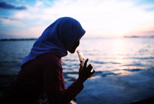 Muslim woman drinks - Muslim woman is drinking a soft drink while sitting on the sea coast and watching the sunset.