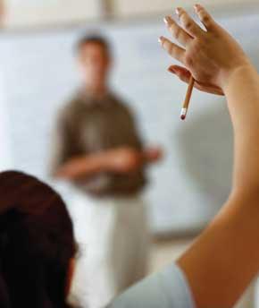 Hand Raised - The photo shows a woman raising her right hand with a pencil and in the background which is in front of the woman, a man which looks to be a teacher or a professor.  This gesture is usually done when one wants to catch a person's attention. In this situation, the woman is trying to catch the professor's attention. She might have a question or perhaps would like to excuse herself for another reason.