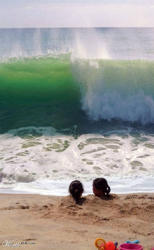 The last second of life! - Picture of two playful girls buried upto their necks in sand and a large wave coming in only a few seconds away, ready to drown them as they look on!